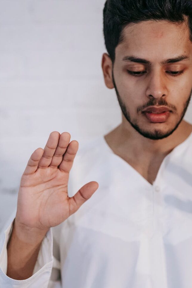 A man wearing traditional attire with raised hand, depicting religious prayer indoors.